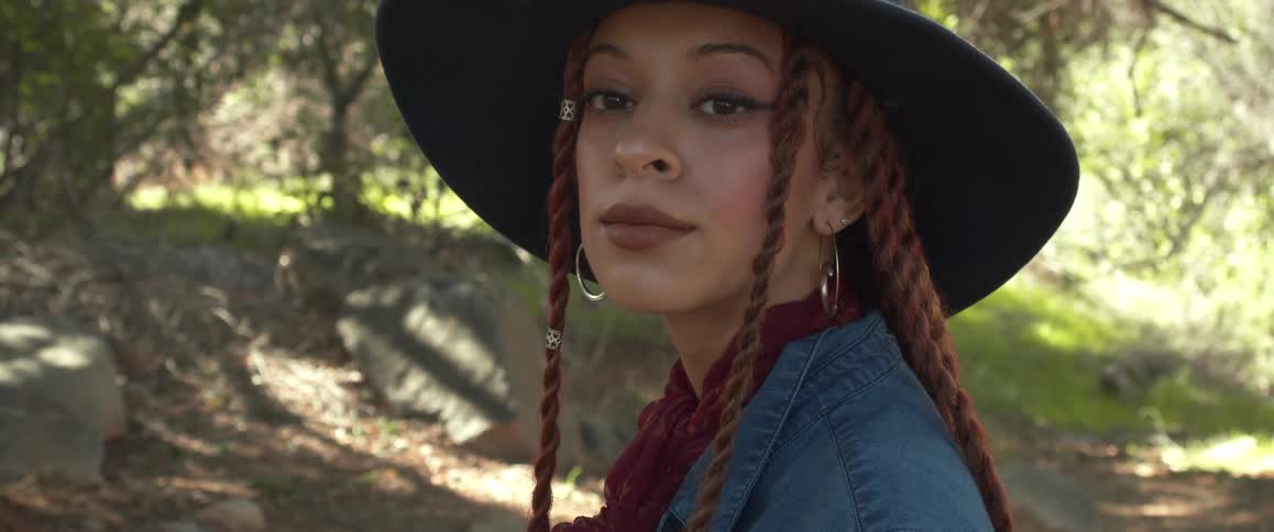 A woman with a hat and earrings standing in a forest with trees and plants in the background