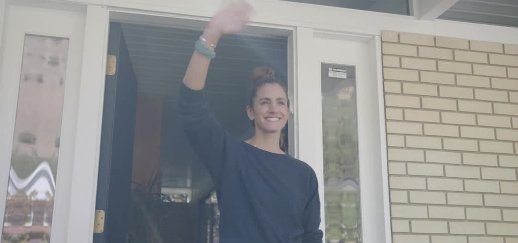 A woman is standing on the front porch of her home while smiling and waving