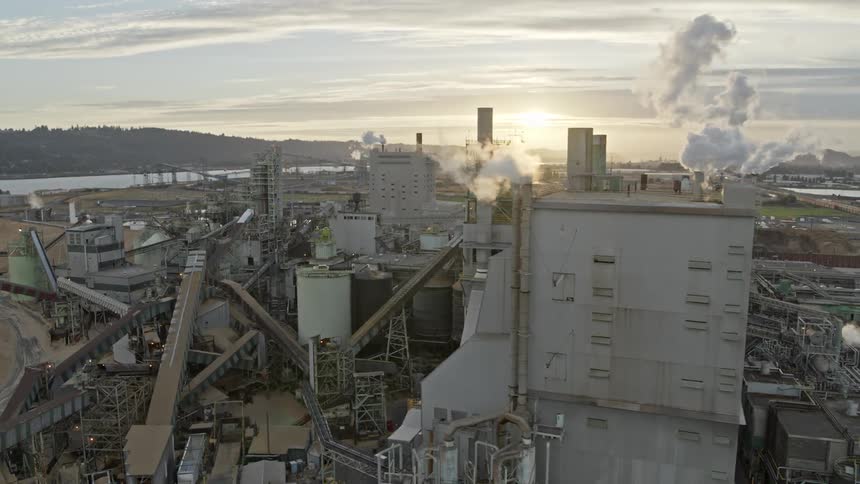 Smoke and steam billow from a dirty gray factory building surrounded by heavy industrial manufacturing facilities