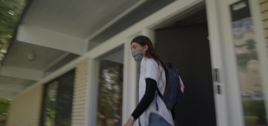 A teenager walks out of their home with a backpack and wearing a facemask and walks down the sidewalk towards the road