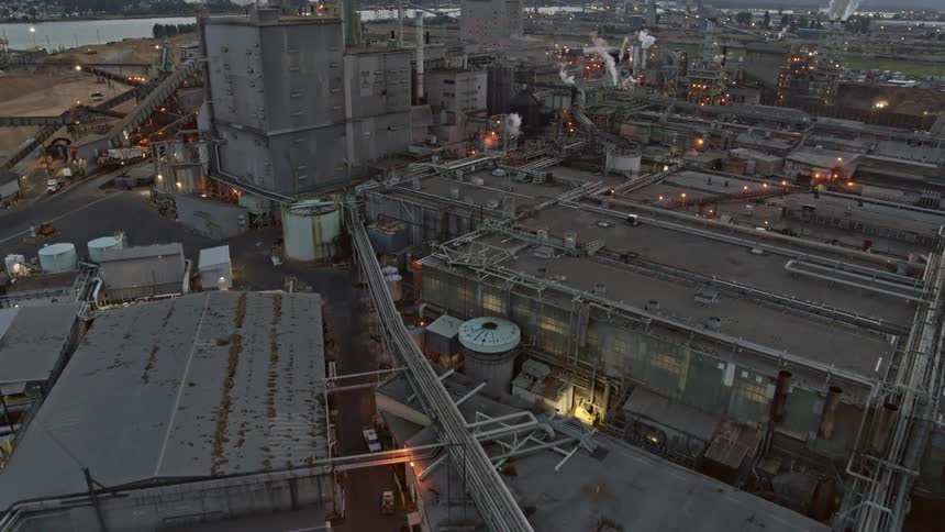 Flying low over a maze of pipes and factory buildings as smoke and steam rise from various stacks