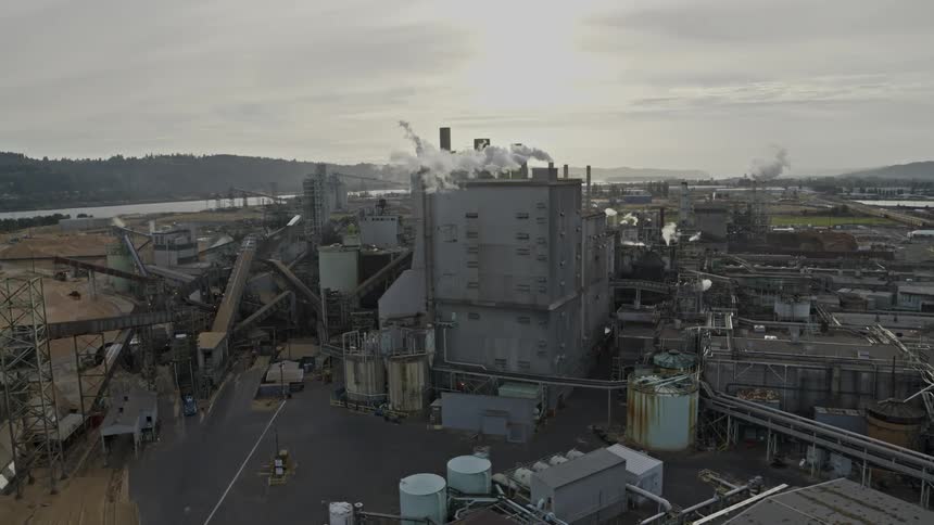 Industrial pollution rises from a large factory complex backlit by the setting sun