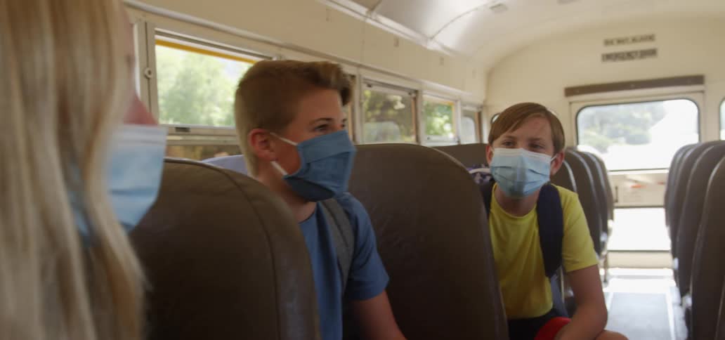 Two boys and a girl wearing face masks sit on a moving school bus talking at daytime