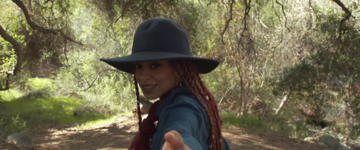 A woman traveling through a woodland during the day looks back to gesture for the camera to follow