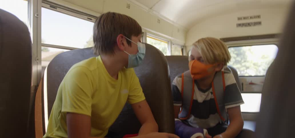 Two boys with face masks sitting on a school bus and talking