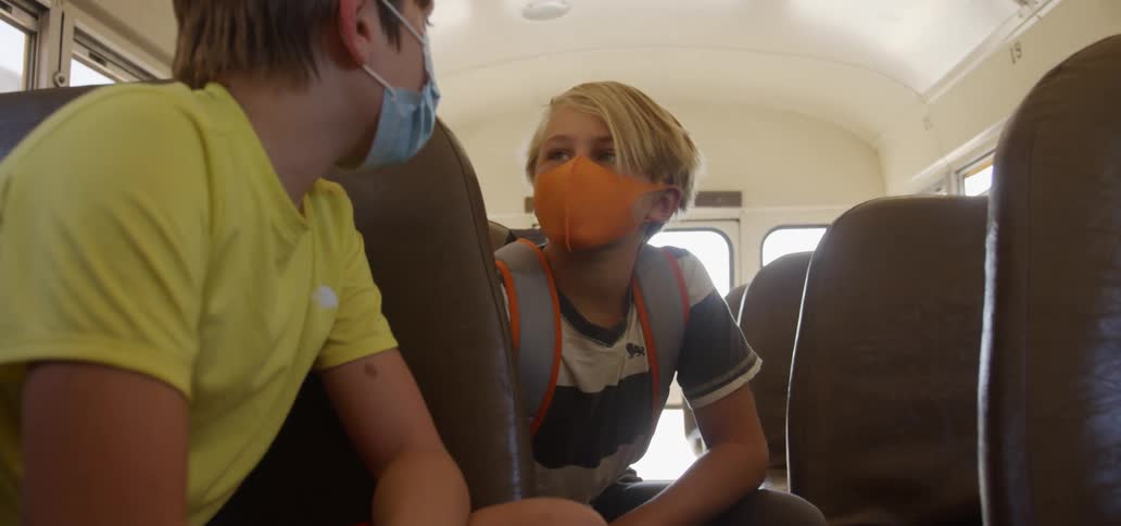 Two boys sit on a moving school bus talking while wearing face masks