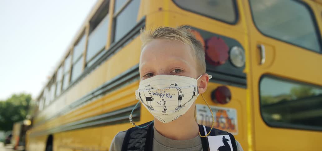 A young boy faces a face mask while standing in front of a school bus parked outside his house