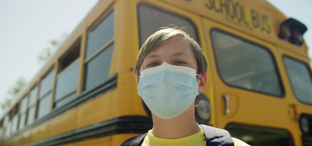 A child wearing a face mask is standing behind a school bus in front of a school during the day