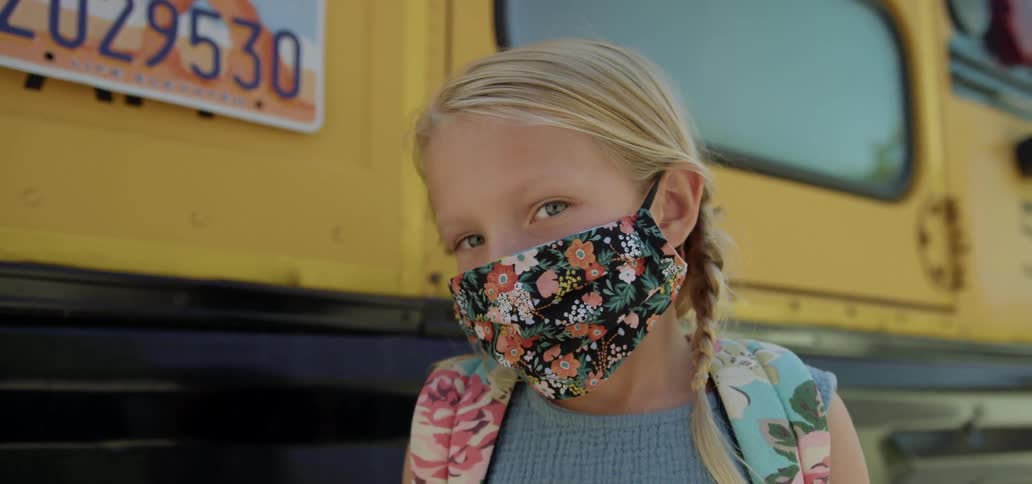 A young girl standing in front of a school bus with a mask on