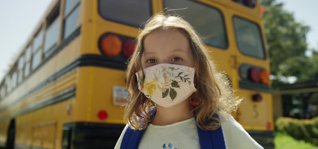A girl with a mask and a backpack standing and staring with a school bus a house trees the sky and clouds in the background