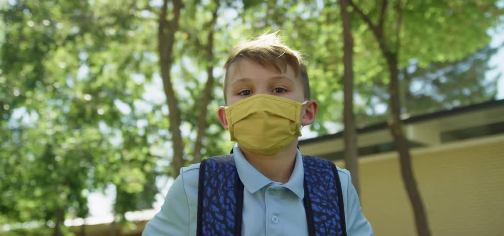 A boy with a backpack and a mask standing and staring with trees a building the sky and clouds in the background