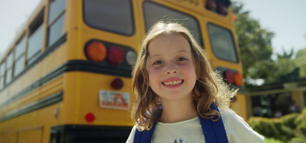 Little girl with a backpack smiles broadly in front of school bus yellow
