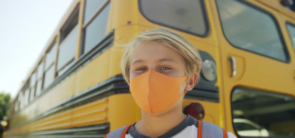A young boy standing in front of a school bus wearing a mask