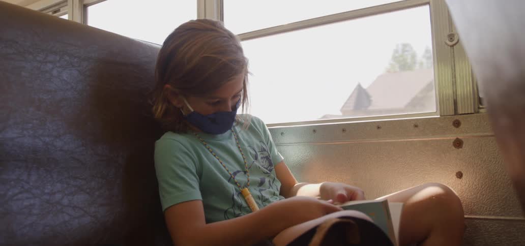 A young girl is riding in a school bus and reading a book while wearing a face mask