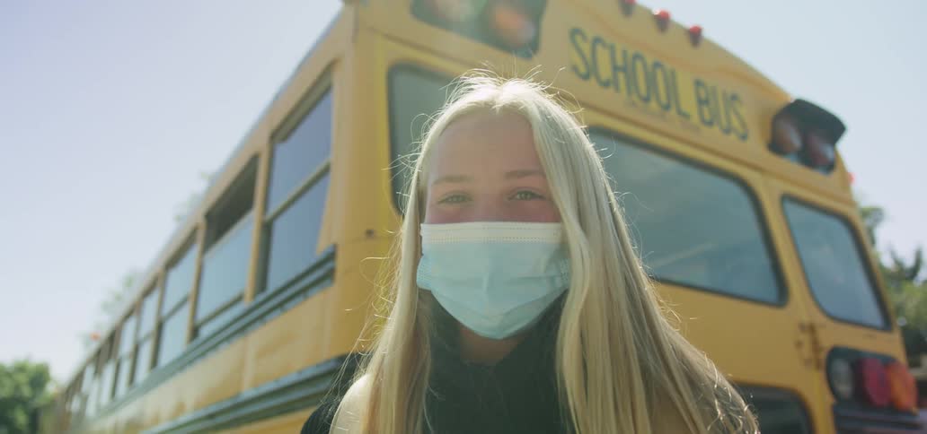 A girl wearing a mask stands in front of a school bus