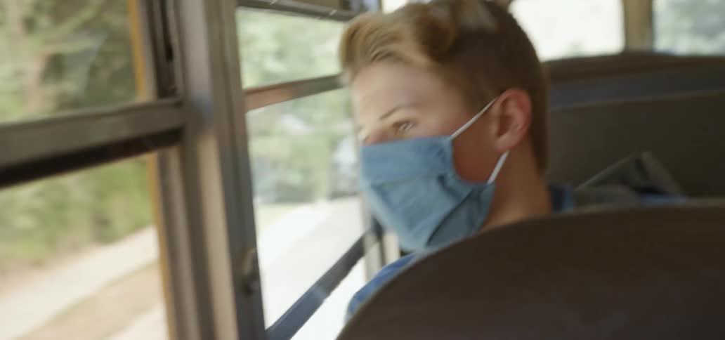 Blonde teenage boy with a blue mask looks out the side windows of the bus