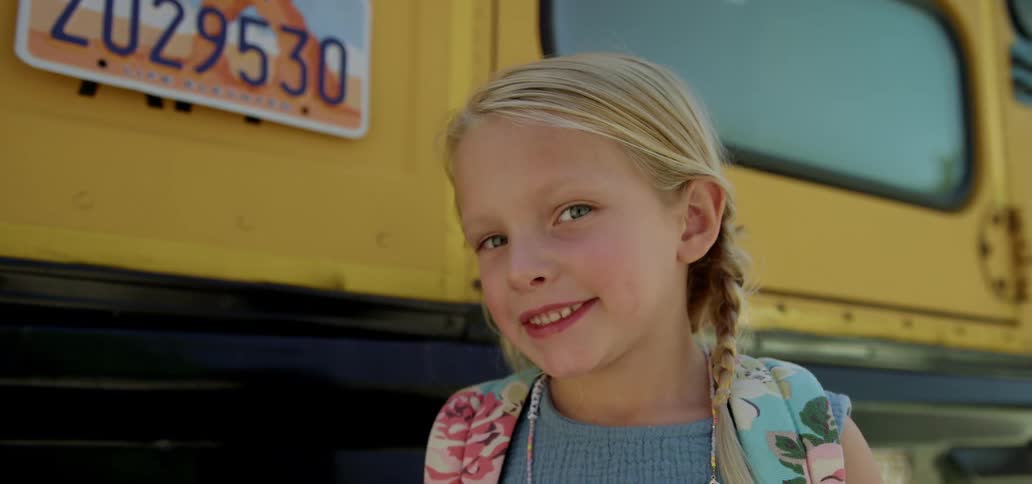 A girl with a backpack standing and smiling with a school bus trees the sky and clouds in the background