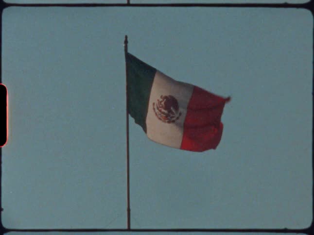 A mexican flag waving in the wind with the sky and clouds in the background