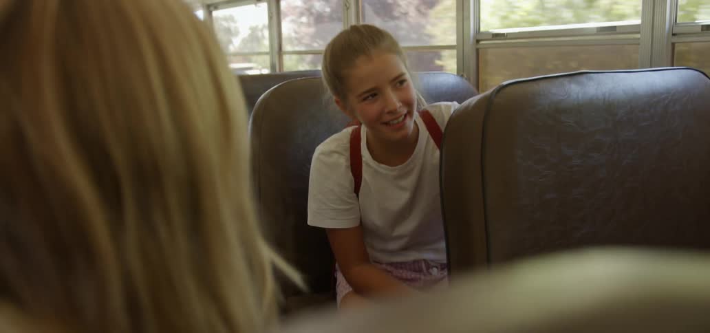 A young girls is sitting in a moving bus talking and laughing to a child across the aisle of the bus during the day