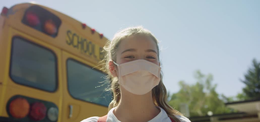 A girl in a medical mask smiles into the camera while standing outdoors behind a school bus