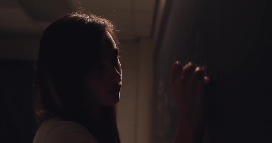 A young woman writes on a chalkboard in very low lighting