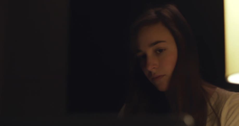 Teenage girl with a long brown hair and a white shirt writes in a notebook in a dark room