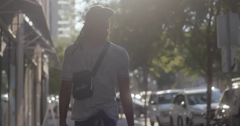 A man with a hat and glasses walking on a city street with trees the sky and clouds in the background