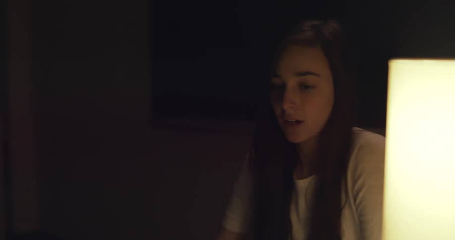 A young woman sitting at a desk turns on a lamp and opens a laptop to begin studying at night