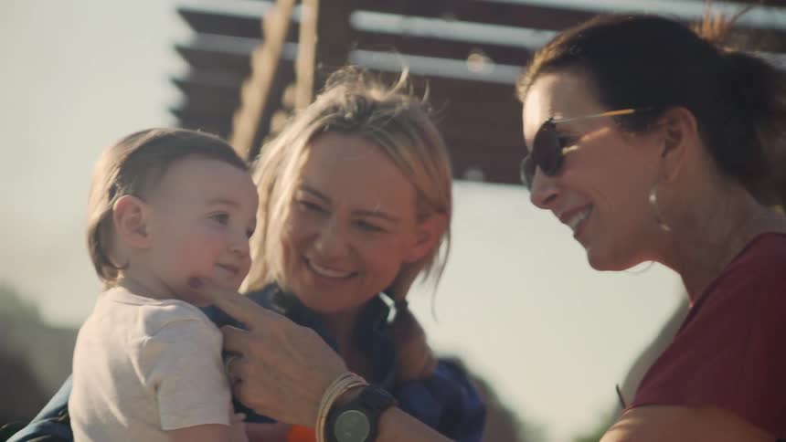 A woman holds her son and smiles as a friend touches him as they stand under a pavilion