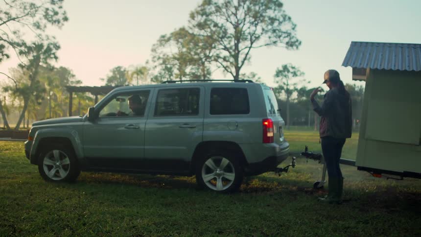 Woman standing in a park helping a man park a car next to a detachable trailer