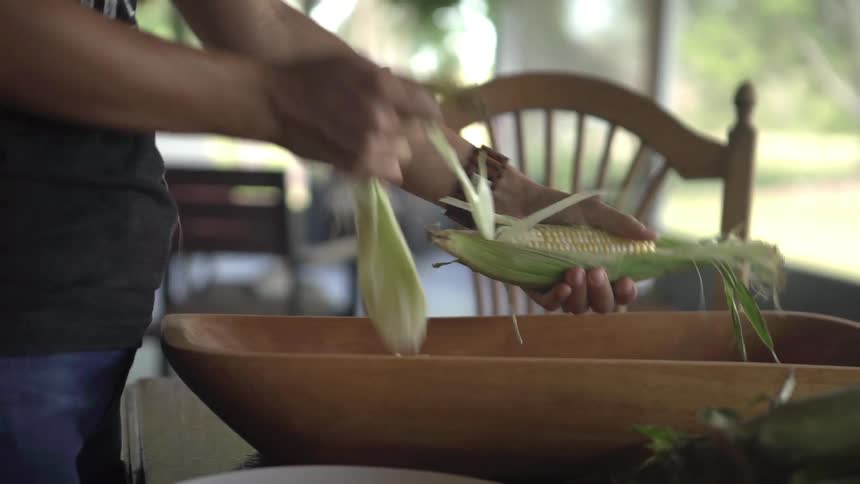 A woman stands at a table shucking corn in a screened in room during the day