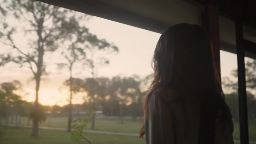 A woman with long dark hair stands on her screened in porch and looks outside