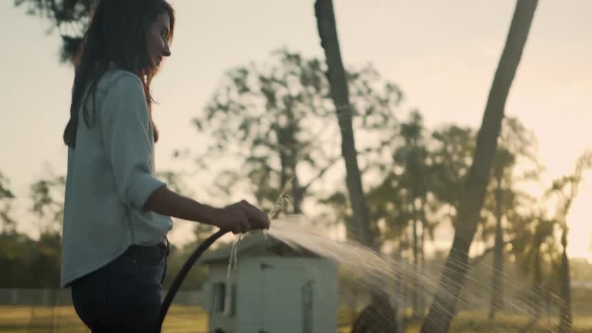 A woman uses a hose to water a plant outside near a garden at dusk