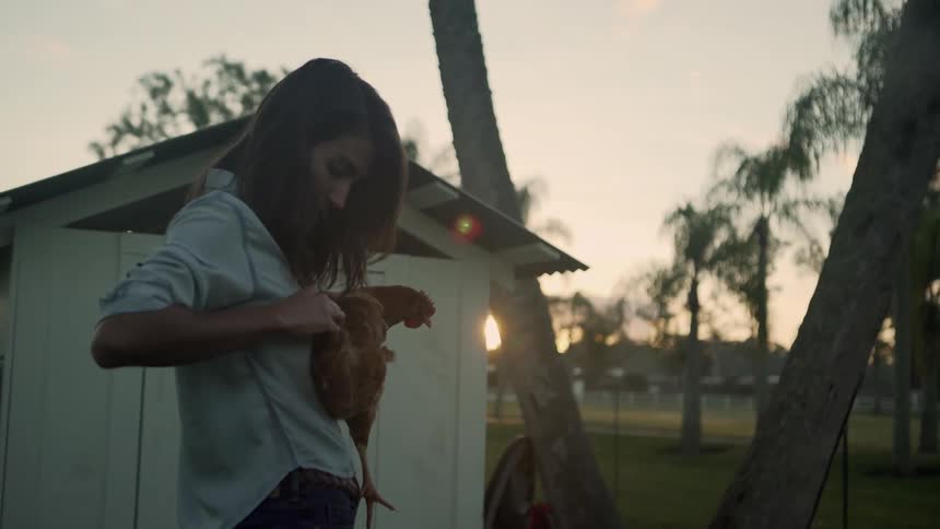 A woman holding a chicken and throwing it on the ground with trees the sky and clouds in the background