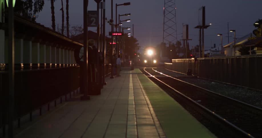 People are waiting on a platform at an outdoor train station as a train approached with bright flashing headlights at night