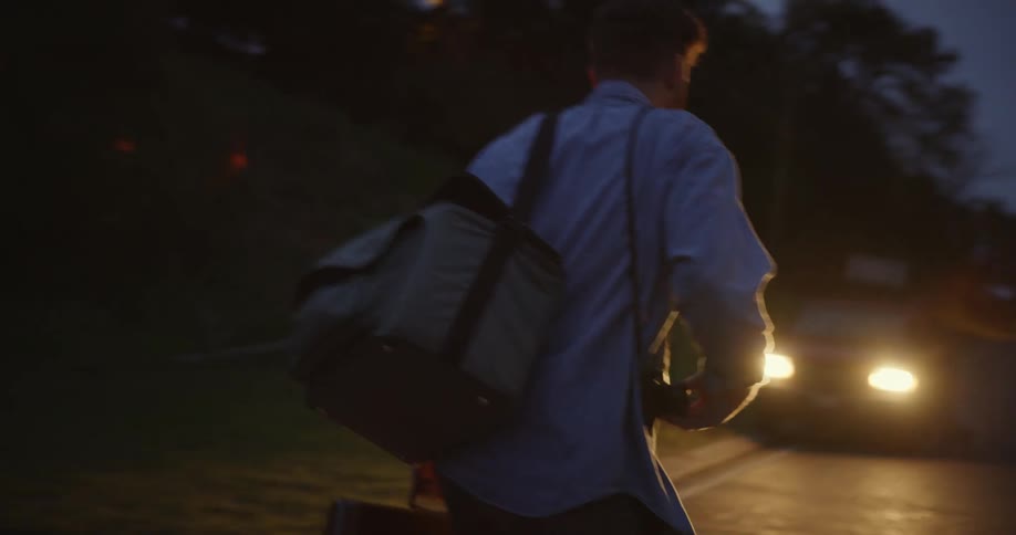 A man carrying baggage heads toward a waiting car with its headlights on at night