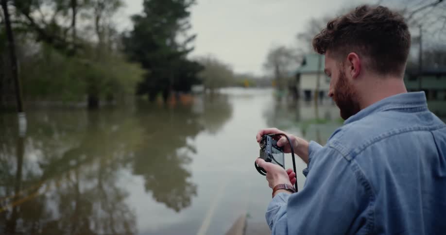 A man with a camera taking pictures of a flooded neighborhood with houses trees the sky and clouds in the background