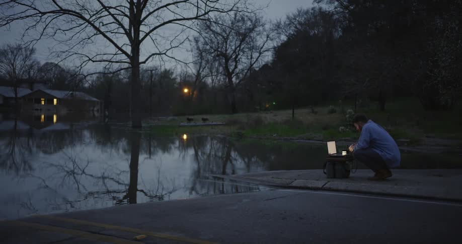 A man knees by a flooded street in a neighborhood and types on a laptop
