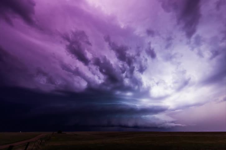 A grassy plain with a dirt road with lightning the sky and clouds above