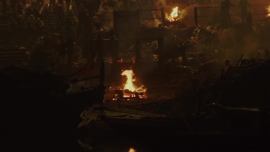 A crowd of people sit in bleachers and stand above a scene where fires are burning
