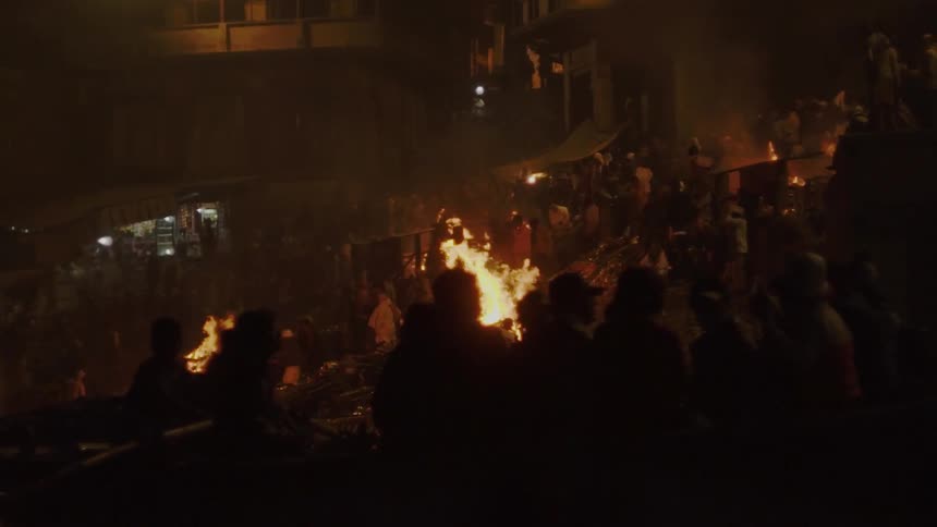 People standing and sitting outside next to large bonfires