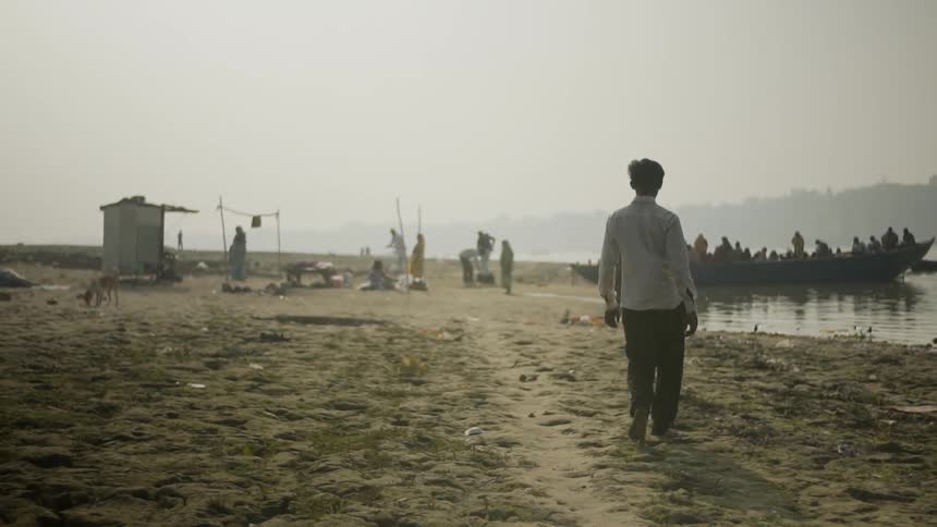 A man walks on the beach toward a large group of people standing in a boat near shacks