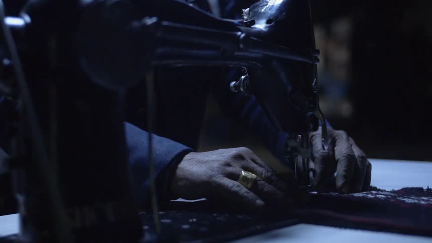 A person sits at a table using a sewing machine in a dimly lit room