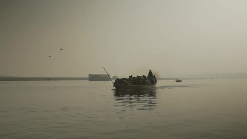 A group of people ride a crowded boat along the coast as birds fly overhead on a foggy day