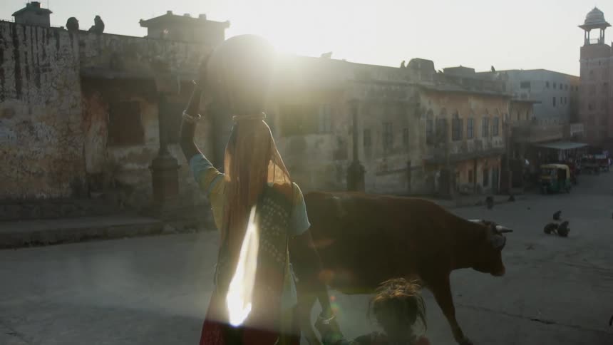 A woman walking on a street with a jug on her head holding a little girls hand with animals on the street with buildings the sky and clouds in the background