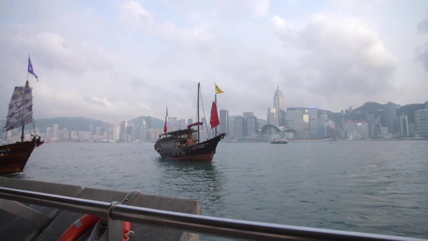 A ferry and two sailboats sit in the ocean across from tall skyscrapers downtown