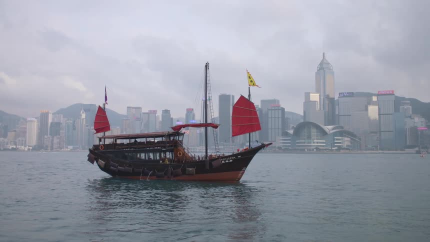 A ship with red sails moves slowly in the ocean as it passes by downtown skyscrapers