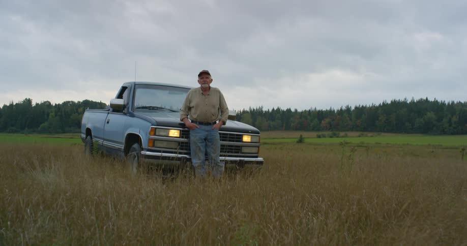 An older man leans against the front of a truck parked on a field at dusk with the headlights on