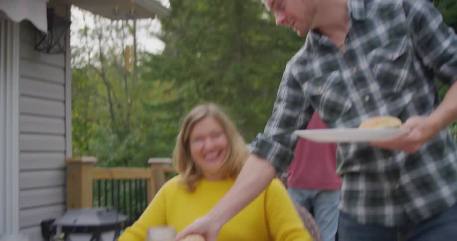 A man brings over a plate of hamburgers to two people sitting at a patio table