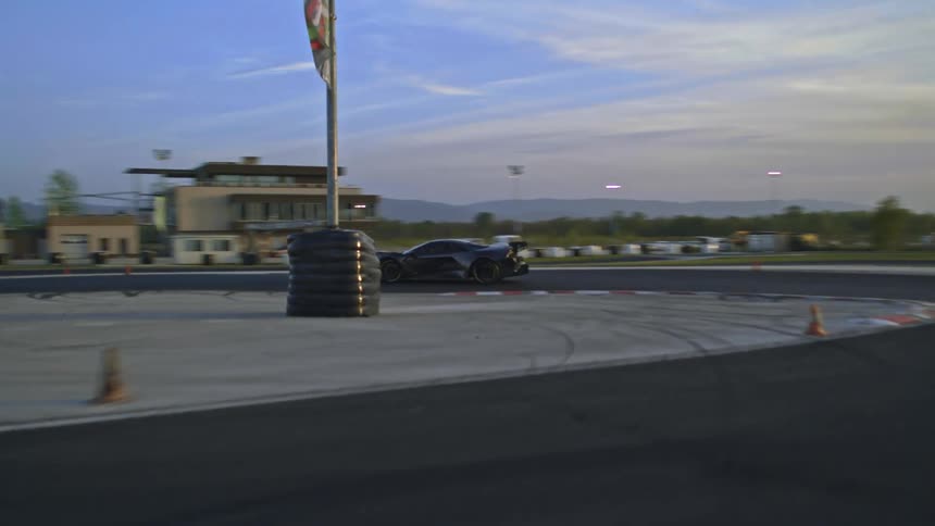A sports car driving on a track with buildings trees the sky and clouds in the background